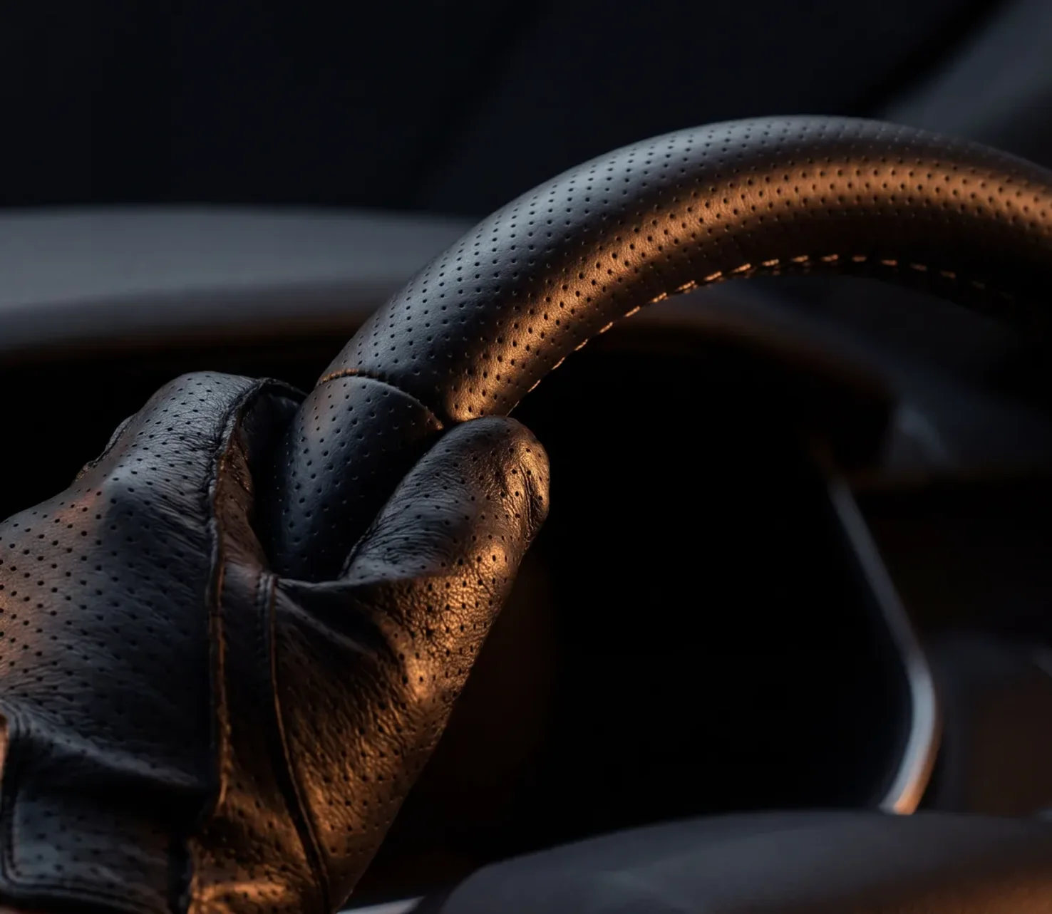 Hand in black perforated leather driving glove gripping the top of a premium luxury SUV steering wheel, warm amber cabin light raking across the leather against deep black negative space